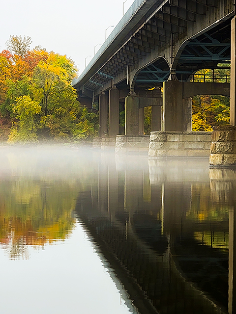 Mist creeps beneath the bridge over the Connecticut River in Suffield, Connecticut, obscuring the start of reflection and the end of shoreline.